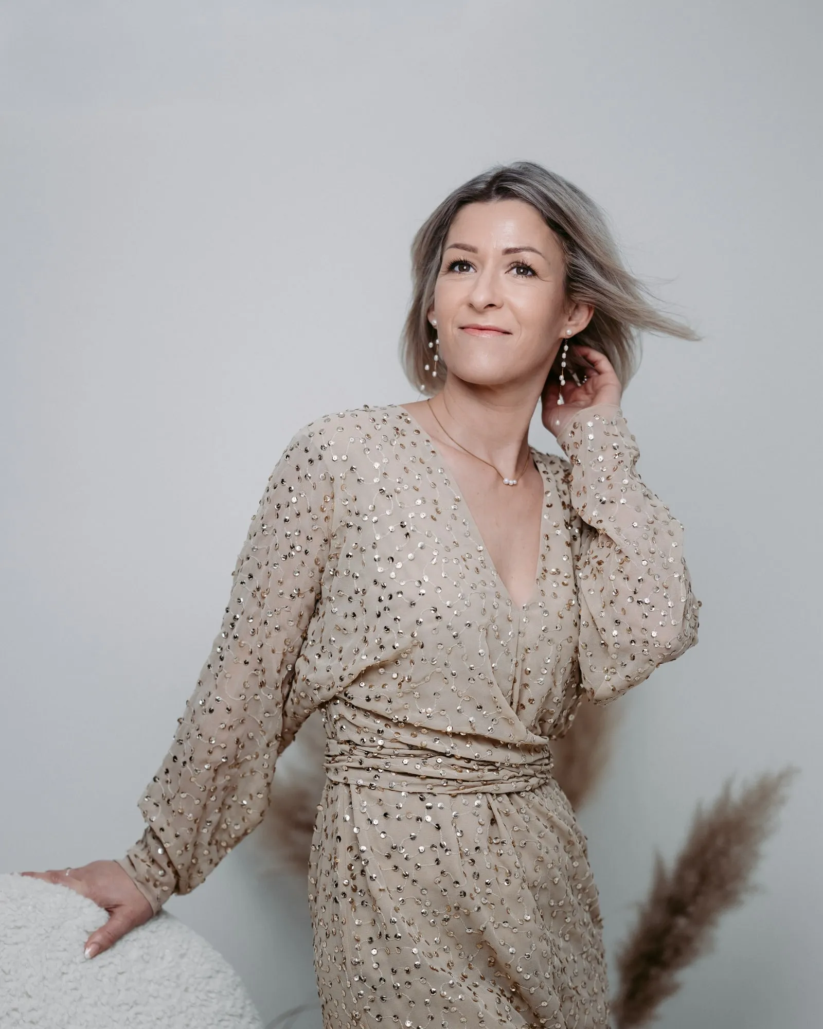 Christina Hohensinn im Pailletten-Kleid, lächelnd an eine helle Lehne gestützt — Visagistin von Beauty by Christina