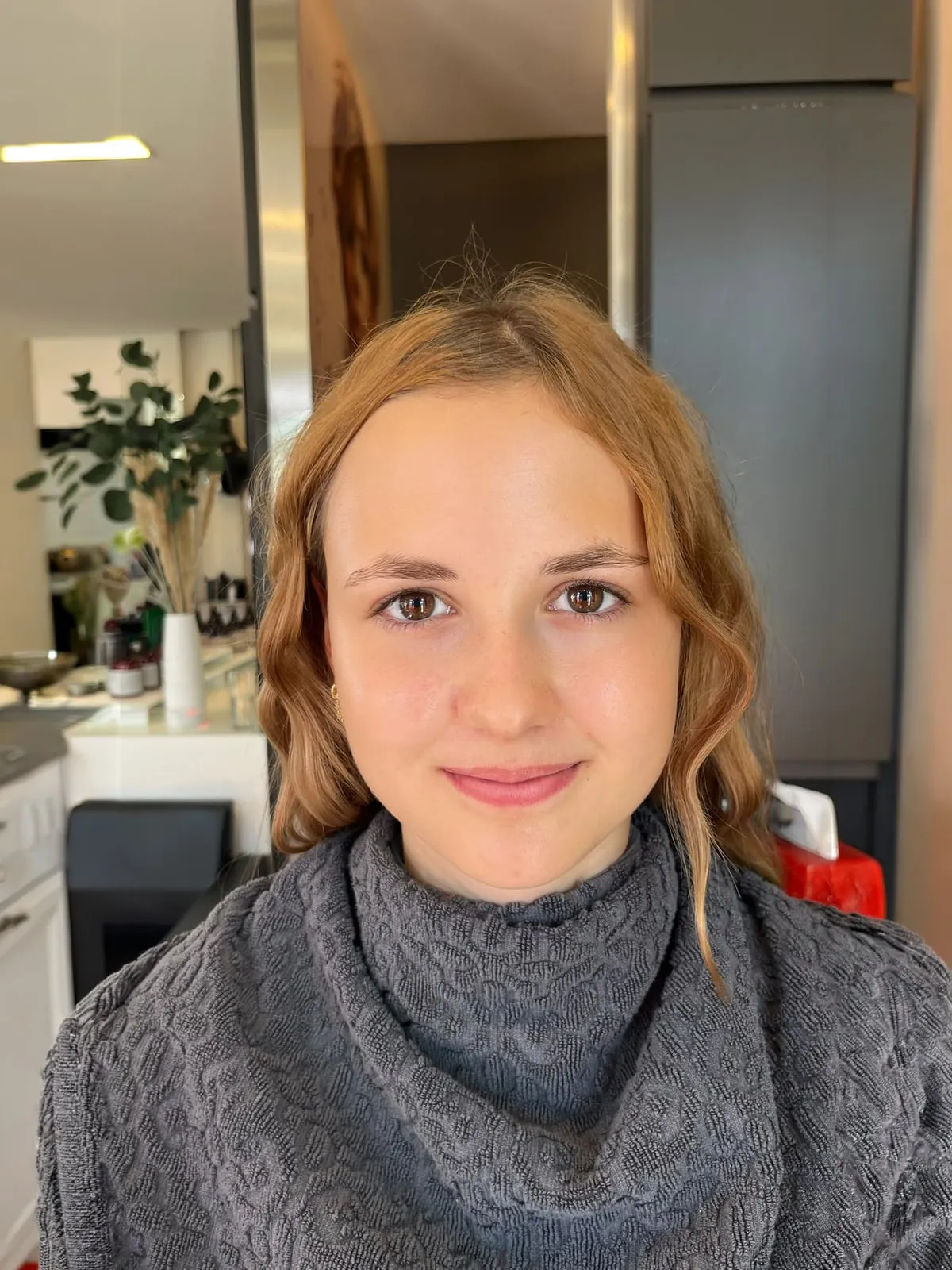 Dieselbe junge Frau ungeschminkt mit rotblonden Wellen und Frisierumhang, vor dem Soft-Glam-Make-up